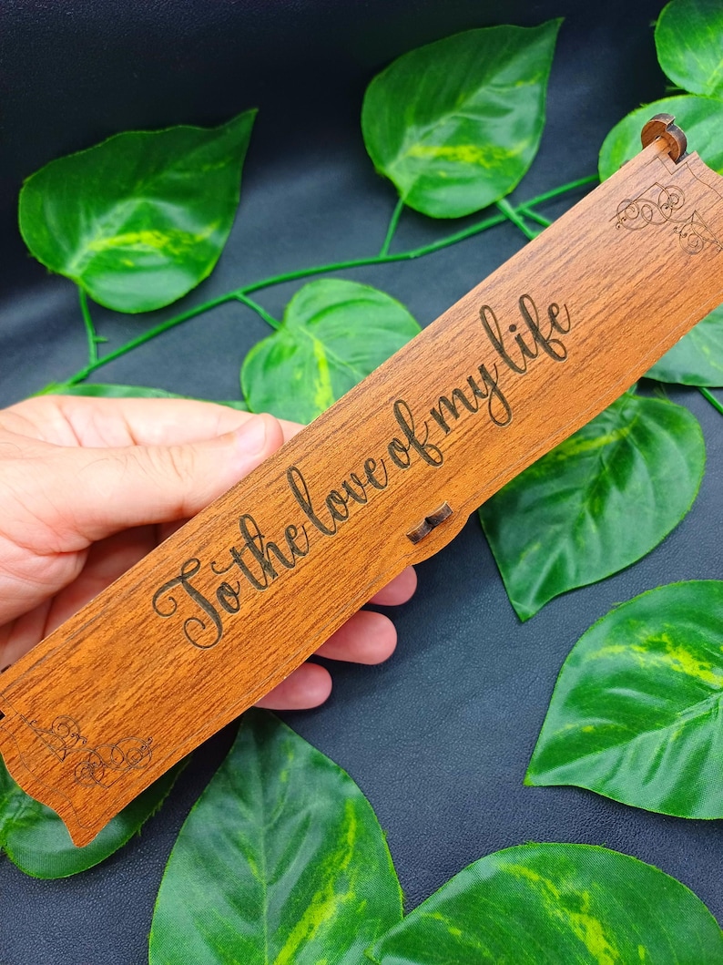 May include: A wooden box with the inscription "To the love of my life" engraved on the top. The box is rectangular, with a natural wood grain finish. Decorative flourishes are etched on the ends. The box is set against a backdrop of green leaves.
