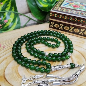 May include: A strand of dark green prayer beads with silver accents and a decorative tassel. The beads are round and evenly spaced. A small, decorated wooden box is in the background.