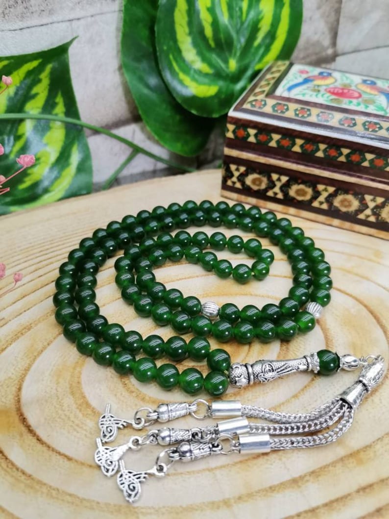 May include: A strand of dark green prayer beads with silver accents and a decorative tassel. The beads are round and evenly spaced. A small, decorated wooden box is in the background.