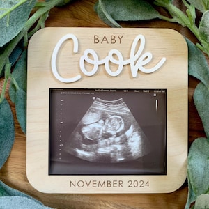 May include: A wooden frame with a white cutout of the name "Baby Cook" and the date "November 2024". The frame holds a black and white ultrasound image.