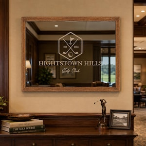 May include: A rectangular wall mirror with a textured brown wooden frame. The glass features a white graphic of crossed golf clubs and a ball inside a hexagon, with the text "EST 2020", "HIGHTSTOWN HILLS", and "Golf Club" printed below.