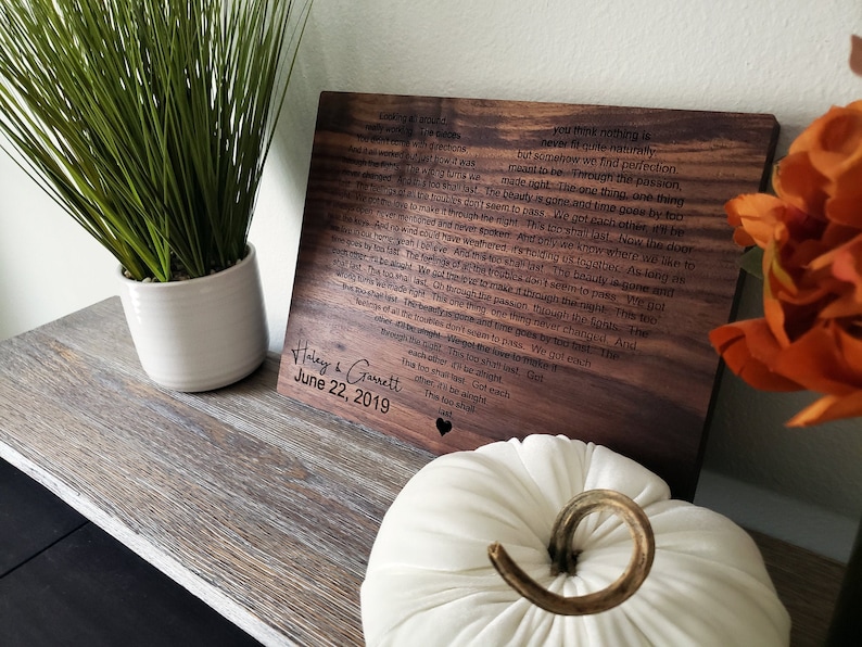 May include: A wooden plaque with a romantic poem engraved on it. The plaque is dark brown and has a heart-shaped cutout at the bottom. The text on the plaque reads: "You think nothing is never quite naturally, but somehow we find perfection.  ...  We got each other. This too shall last."