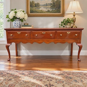 May include: A long wooden console table with three drawers, each featuring a brass keyhole plate and two round knobs. The table has a scalloped apron and four curved legs. It is topped with a blue and white vase of white hydrangeas.