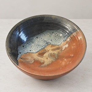 May include: A round ceramic bowl featuring a reactive glaze finish. The interior displays a blend of deep black, speckled light blue, and warm terracotta orange tones, with visible concentric ridges along the inner surface of the bowl.