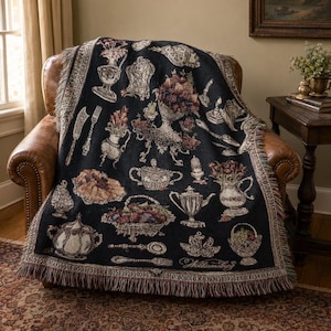 May include: A black woven throw blanket featuring a repeating pattern of vintage-style silver tableware, including teapots, pitchers, bowls of fruit, forks, and spoons, with a decorative fringed edge along the bottom and sides of the fabric.