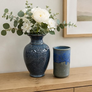 Puede incluir: Un jarrón de cerámica azul oscuro moteado con borde acampanado que contiene flores blancas y tallos de eucalipto verdes, junto a un recipiente cilíndrico de cerámica con un acabado de esmalte reactivo azul y gris sobre una superficie de madera.