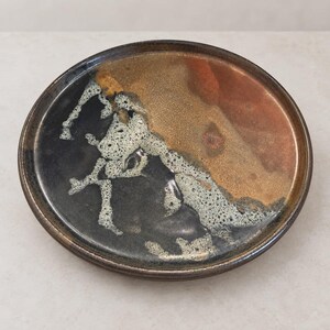 May include: A round ceramic plate featuring a multi-colored glaze with a glossy finish. The surface displays a blend of deep black, metallic bronze, and copper tones, accented by a textured, speckled white and gray reactive glaze pattern.