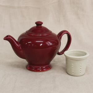 Peut inclure: Une théière en céramique bordeaux profond avec un couvercle assorti, une anse incurvée et un bec verseur, accompagnée d'une infuseur à thé cylindrique blanc cassé séparé doté de petits trous perforés sur les côtés.