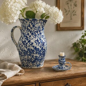 May include: A ceramic pitcher and a matching candle holder, both featuring a blue and white speckled pattern. The pitcher is filled with large white hydrangea flowers, and the candle holder contains a small, plain white pillar candle.