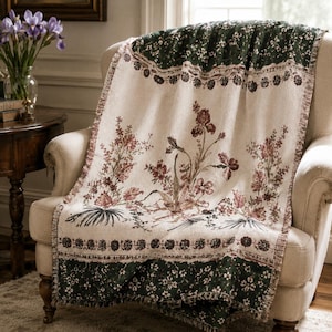 May include: A woven throw blanket featuring a cream-colored center with a botanical print of reddish-brown flowers and green foliage. The blanket has dark green borders at the top and bottom decorated with a small white floral pattern.