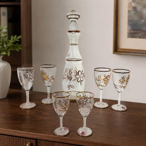 May include: A clear glass decanter with a matching stopper and six stemmed glasses, all featuring a delicate gold floral pattern and gold-rimmed edges. The set includes a tall, slender decanter and six small glasses with thin stems and circular bases.