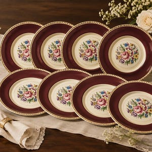 May include: A set of eight round dinner plates featuring a deep burgundy rim with a gold-toned beaded edge and a cream-colored center decorated with a colorful floral bouquet of pink roses, blue flowers, and green leaves.
