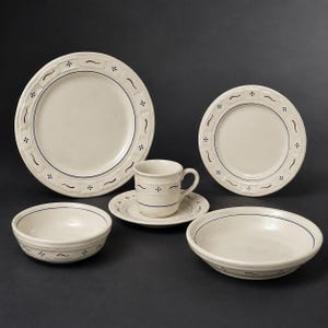 May include: A cream-coloured ceramic dinnerware set featuring a dark blue line pattern along the rims. The set includes a large dinner plate, a smaller salad plate, a bowl, a wide shallow bowl, and a coffee cup resting on a matching saucer.