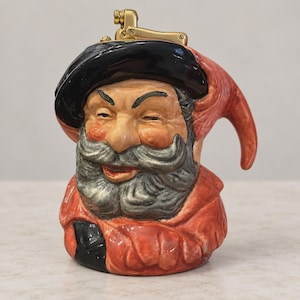 Puede incluir: Un encendedor de mesa de cerámica con forma de busto de una figura barbuda que lleva una boina negra y una prenda roja. La figura tiene bigote y barba grises, con un mecanismo de encendedor de metal dorado fijado sobre la boina negra.
