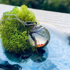 Peut inclure: Un collier en chaîne argentée avec un pendentif en verre transparent. Le pendentif contient un petit palmier en résine rouge et blanc. Le pendentif est posé sur une tranche d'agate bleue et blanche.
