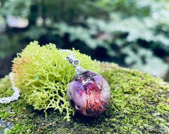 May include: A pendant necklace with a clear resin orb, containing dried purple and red flowers. The silver chain supports the orb, which sits on green moss and lichen. The necklace is a unique, nature-inspired piece.
