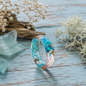 Può includere: Un anello in resina trasparente con scaglie iridescenti blu e turchesi incorporate e accenti metallici color rame. La fascia presenta una finitura liscia e lucida con un profilo arrotondato, mostrando un aspetto vivace e riflettente multicolore.