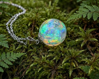 May include: A spherical pendant necklace featuring a clear resin orb filled with iridescent, multi-colored opal-like flakes that shimmer with green, blue, and orange hues. The orb is attached to a silver-toned metal bail and a delicate silver chain.