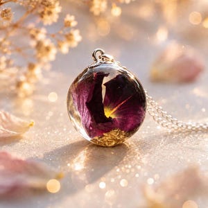 May include: A spherical resin pendant necklace featuring a deep purple flower petal and gold leaf flakes suspended inside the clear material. The pendant is attached to a delicate silver-toned metal chain, creating a clear, circular jewellery piece.