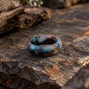May include: A smooth, rounded ring featuring a swirling blend of deep blue and translucent white resin, accented with embedded metallic copper flakes throughout the band, creating a marbled appearance with a polished, glossy finish.