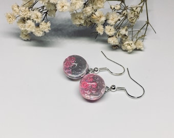 May include: A pair of pink and clear glass earrings with silver hooks. The earrings are round and have a textured surface.