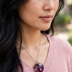 May include: A silver chain necklace featuring a spherical clear resin pendant that encapsulates a deep purple flower and small gold-coloured flecks. The pendant is attached to the chain with a decorative silver-toned metal cap at the top.