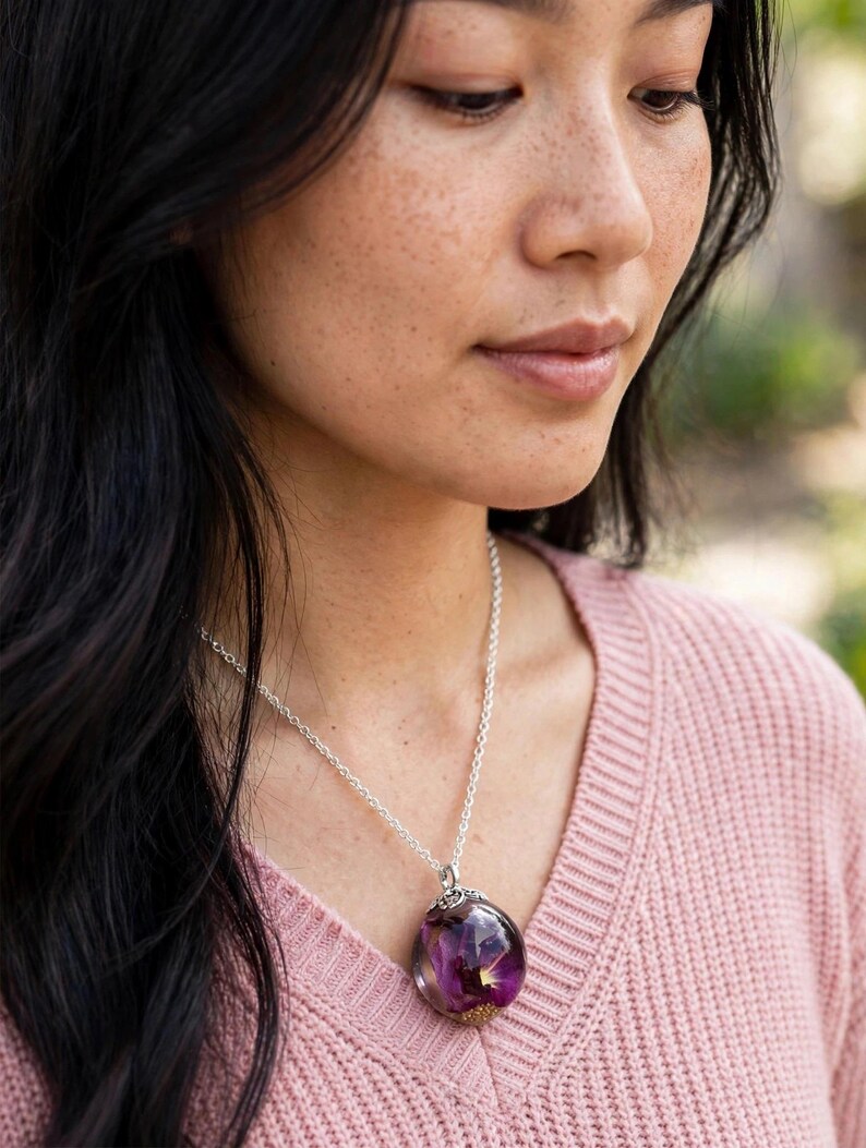 May include: A silver chain necklace featuring a spherical clear resin pendant that encapsulates a deep purple flower and small gold-coloured flecks. The pendant is attached to the chain with a decorative silver-toned metal cap at the top.