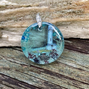May include: A round, clear resin pendant featuring a lighthouse and sea turtles. The pendant has a silver bail with clear rhinestones.