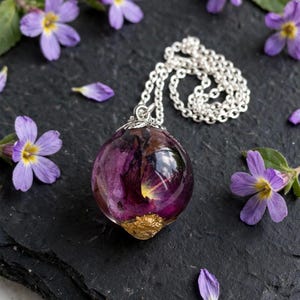 May include: A spherical resin pendant necklace featuring a deep purple flower encased within clear material, accented with a textured gold-coloured base. The pendant hangs from a silver-toned metal chain, showcasing a smooth, polished finish.