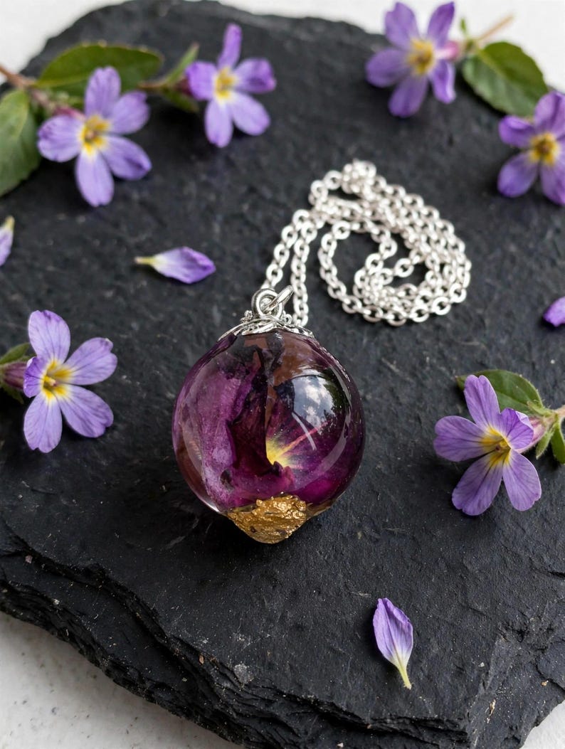 May include: A spherical resin pendant necklace featuring a deep purple flower encased within clear material, accented with a textured gold-coloured base. The pendant hangs from a silver-toned metal chain, showcasing a smooth, polished finish.