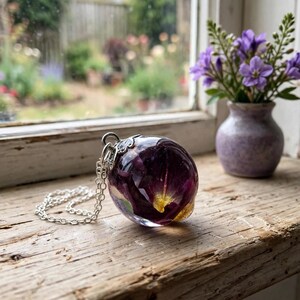 May include: A spherical resin pendant necklace featuring embedded deep purple and yellow flower petals. The clear, polished resin orb is attached to a silver-toned metal bail and a delicate silver-toned link chain, showcasing organic floral elements.
