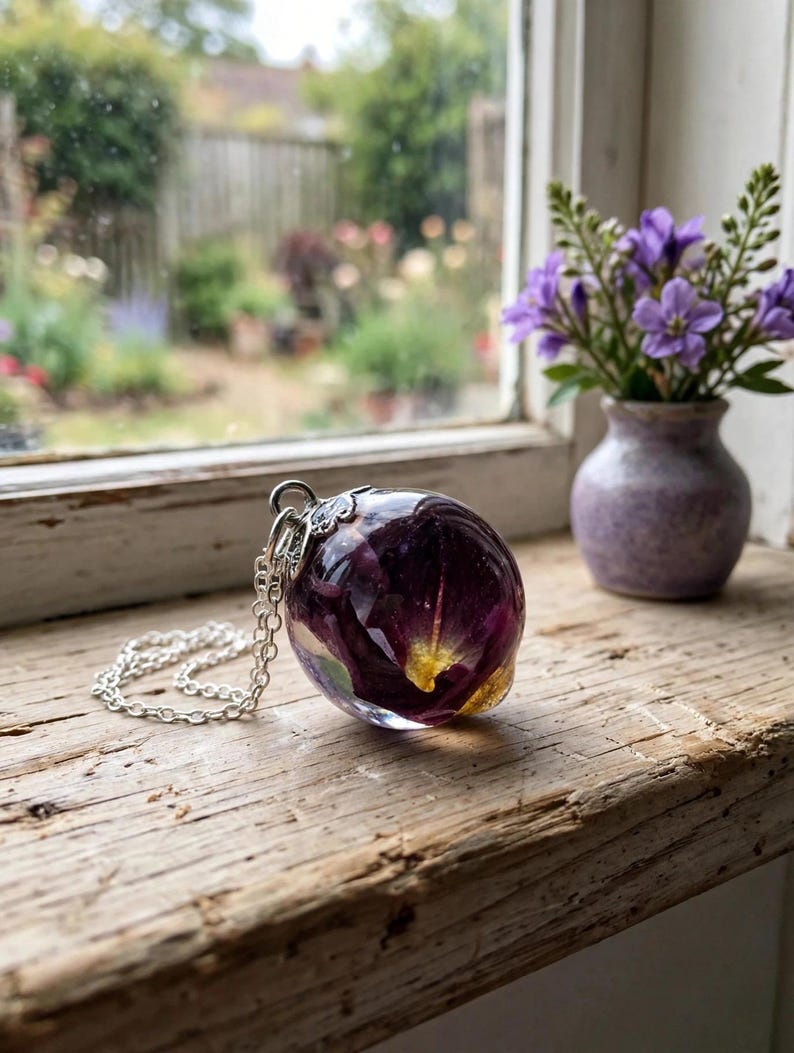 May include: A spherical resin pendant necklace featuring embedded deep purple and yellow flower petals. The clear, polished resin orb is attached to a silver-toned metal bail and a delicate silver-toned link chain, showcasing organic floral elements.