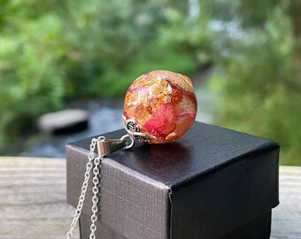 May include: A silver chain necklace with a pendant containing dried red and gold floral elements encased in a clear orb. The pendant is presented on a black box, with a blurred natural backdrop.