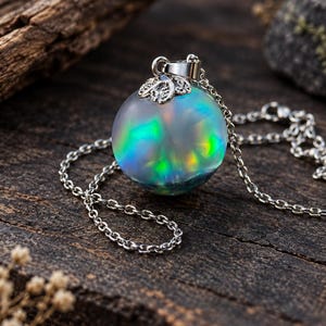 May include: A spherical pendant necklace featuring a translucent orb with iridescent green and blue light reflections. The orb is topped with a silver-toned metal cap with a leaf-like design and is attached to a delicate silver-toned metal chain.
