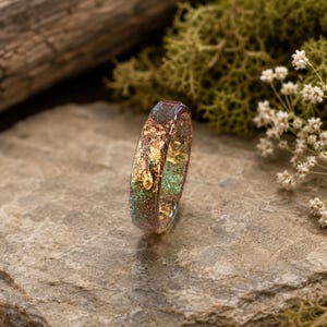 Op de afbeelding: Een ronde ring van doorschijnende hars met ingebedde bladgoudvlokken en fijne groene glitterdeeltjes. De ring heeft een gladde, gepolijste afwerking met een verloop van donkere tinten naar een glinsterende groene kleur.