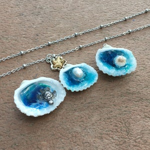 May include: A silver chain necklace with three shell charms. Each shell charm has a blue and white resin ocean scene inside. One shell has a small turtle, one has a pearl, and one has a gold starfish.