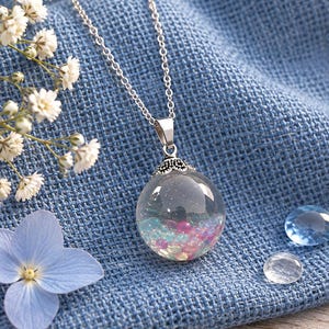 May include: A silver-toned necklace featuring a clear, spherical glass pendant filled with small, iridescent pink and blue beads. The pendant is topped with a decorative silver-toned metal cap and hangs from a delicate silver-toned chain.