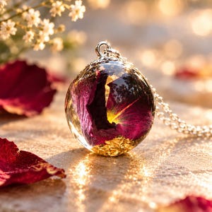 May include: A spherical pendant necklace featuring a deep purple rose petal encased in clear resin with gold leaf flakes at the base. The pendant is attached to a silver-toned metal chain, capturing light with a reflective, glossy finish.