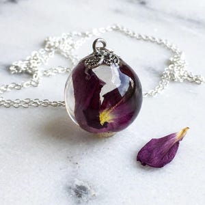 May include: A spherical resin pendant necklace featuring a deep purple rose petal suspended inside clear resin. The pendant is topped with a decorative silver-toned metal cap and hangs from a delicate silver-toned chain. A single loose purple petal lies nearby.