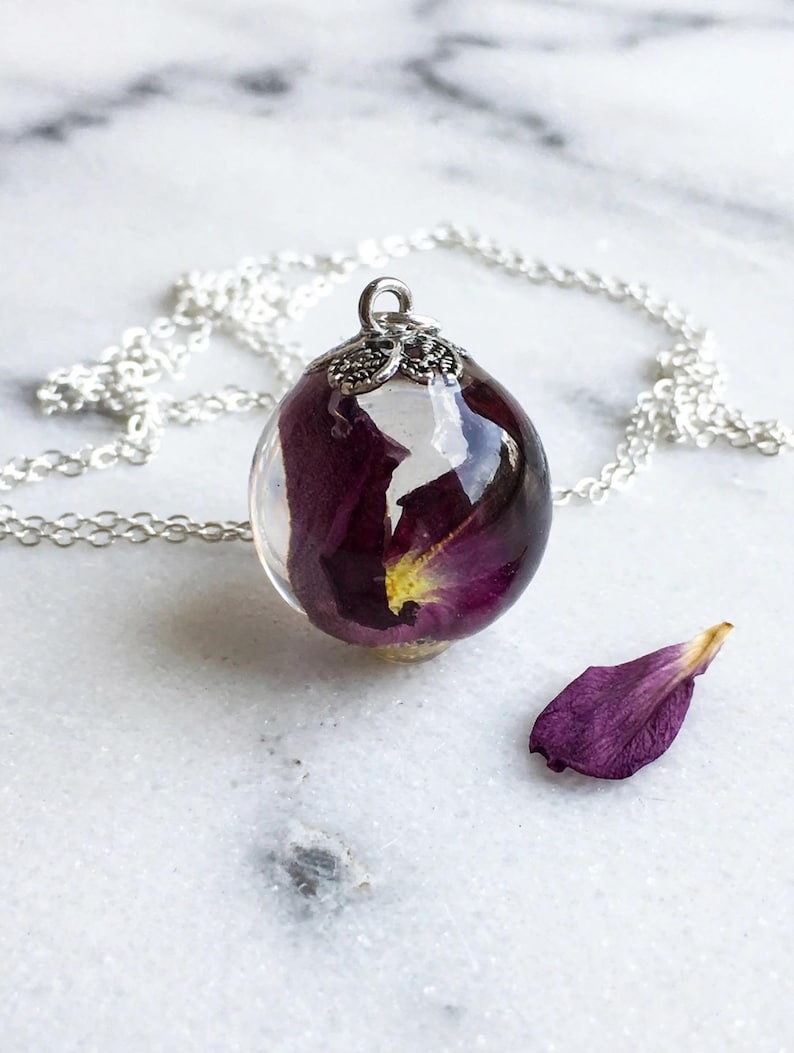 May include: A spherical resin pendant necklace featuring a deep purple rose petal suspended inside clear resin. The pendant is topped with a decorative silver-toned metal cap and hangs from a delicate silver-toned chain. A single loose purple petal lies nearby.