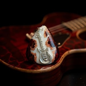 May include: A guitar pick pendant with a silver electric guitar design. The pendant is set against a white background with copper-colored accents. The pick is resting on a red and brown guitar.