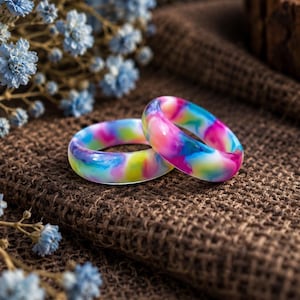 May include: Two smooth, rounded rings featuring a vibrant, swirling marbled pattern of bright pink, electric blue, sunny yellow, and white colors. The rings have a glossy, polished finish and are positioned together, showcasing their colorful, blended resin-like appearance.