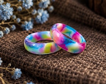 Puede incluir: Dos anillos lisos y redondeados con un vibrante patrón de remolinos jaspeados en colores rosa brillante, azul eléctrico, amarillo soleado y blanco. Los anillos tienen un acabado pulido y brillante y están colocados juntos, mostrando su aspecto colorido y mezclado.