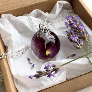 May include: A spherical resin pendant necklace containing a deep purple flower petal, suspended from a silver-toned metal chain. The pendant features a decorative silver-toned cap and is presented with sprigs of dried lavender flowers.