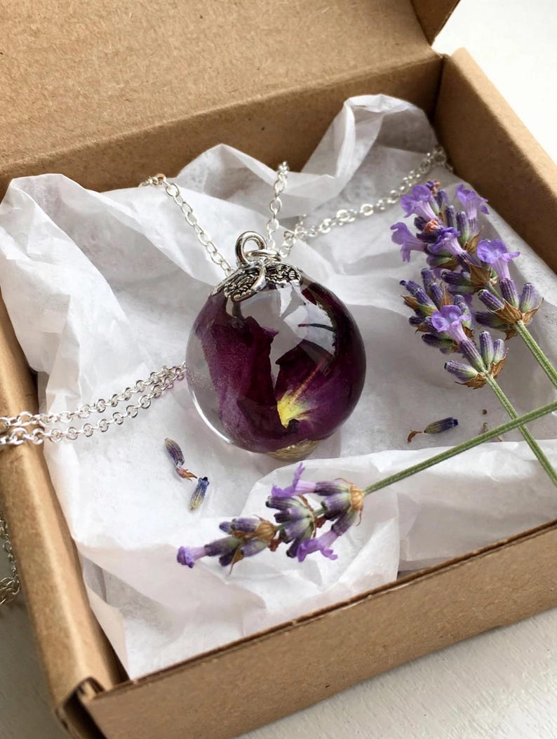May include: A spherical resin pendant necklace containing a deep purple flower petal, suspended from a silver-toned metal chain. The pendant features a decorative silver-toned cap and is presented with sprigs of dried lavender flowers.