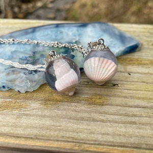 May include: Two silver necklaces with clear glass pendants containing seashells. The shells are a light pink color.