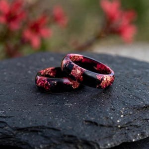 May include: Two rings crafted from a dark, opaque material embedded with shimmering, metallic red foil flakes. The rings feature a smooth, rounded band profile with a glossy finish, showcasing the contrast between the deep black base and the vibrant red accents.