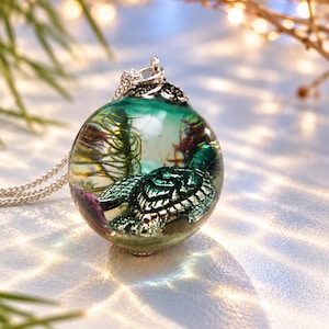 May include: A spherical resin pendant necklace featuring a miniature metallic sea turtle figurine submerged within a clear, teal-tinted environment. The pendant includes delicate green aquatic plant elements and is attached to a silver-toned chain.
