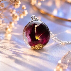 May include: A spherical resin pendant necklace featuring a deep purple flower petal encased within clear resin, accented with gold leaf flakes at the base. The pendant is attached to a silver-toned metal bail and a delicate silver-toned chain.