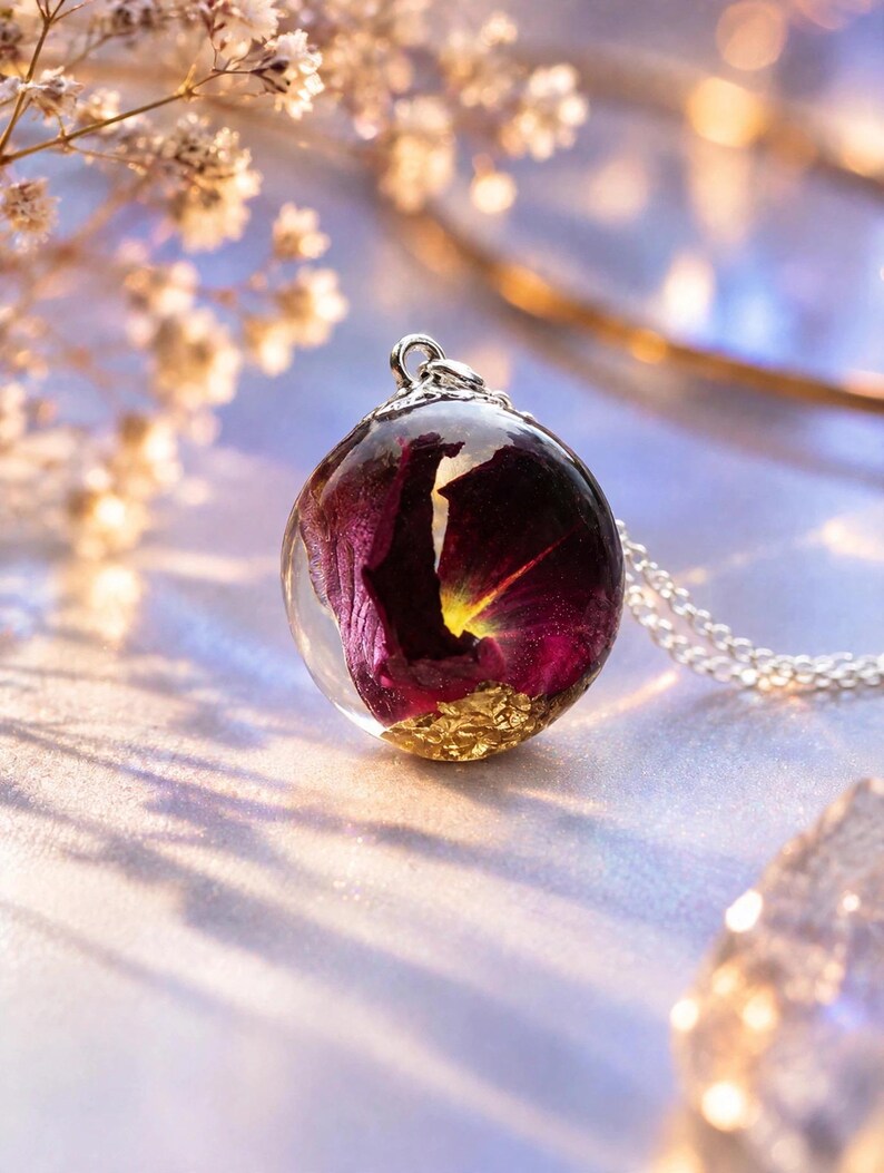 May include: A spherical resin pendant necklace featuring a deep purple flower petal encased within clear resin, accented with gold leaf flakes at the base. The pendant is attached to a silver-toned metal bail and a delicate silver-toned chain.
